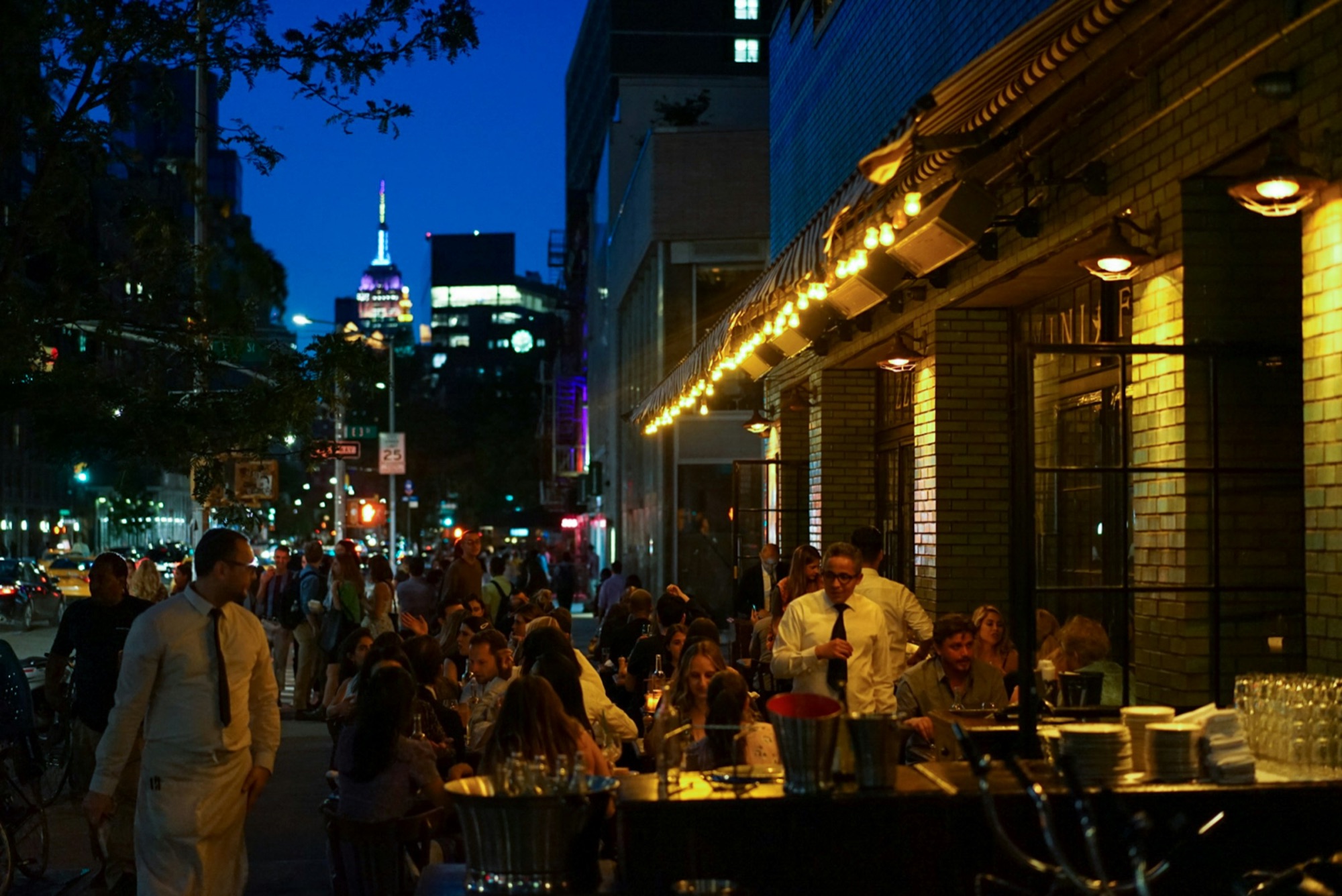 Diners fill a string-lit sidewalk restaurant at night with the Empire State Building glowing above
