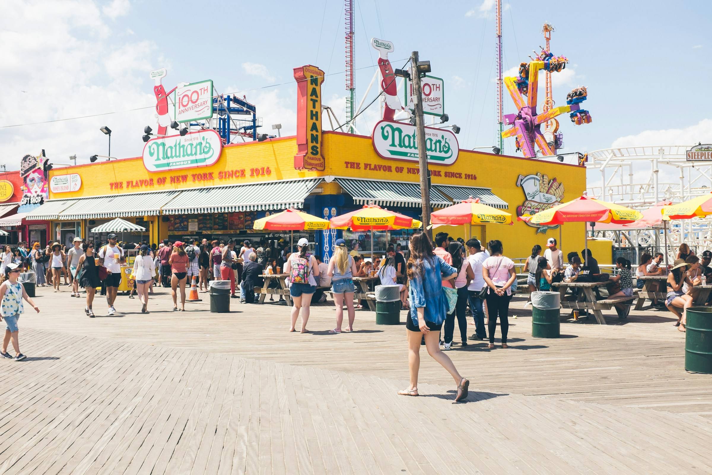 A summer crowd on the Coney Island boardwalk outside Nathan's Famous, with umbrellas, picnic tables and rides behind