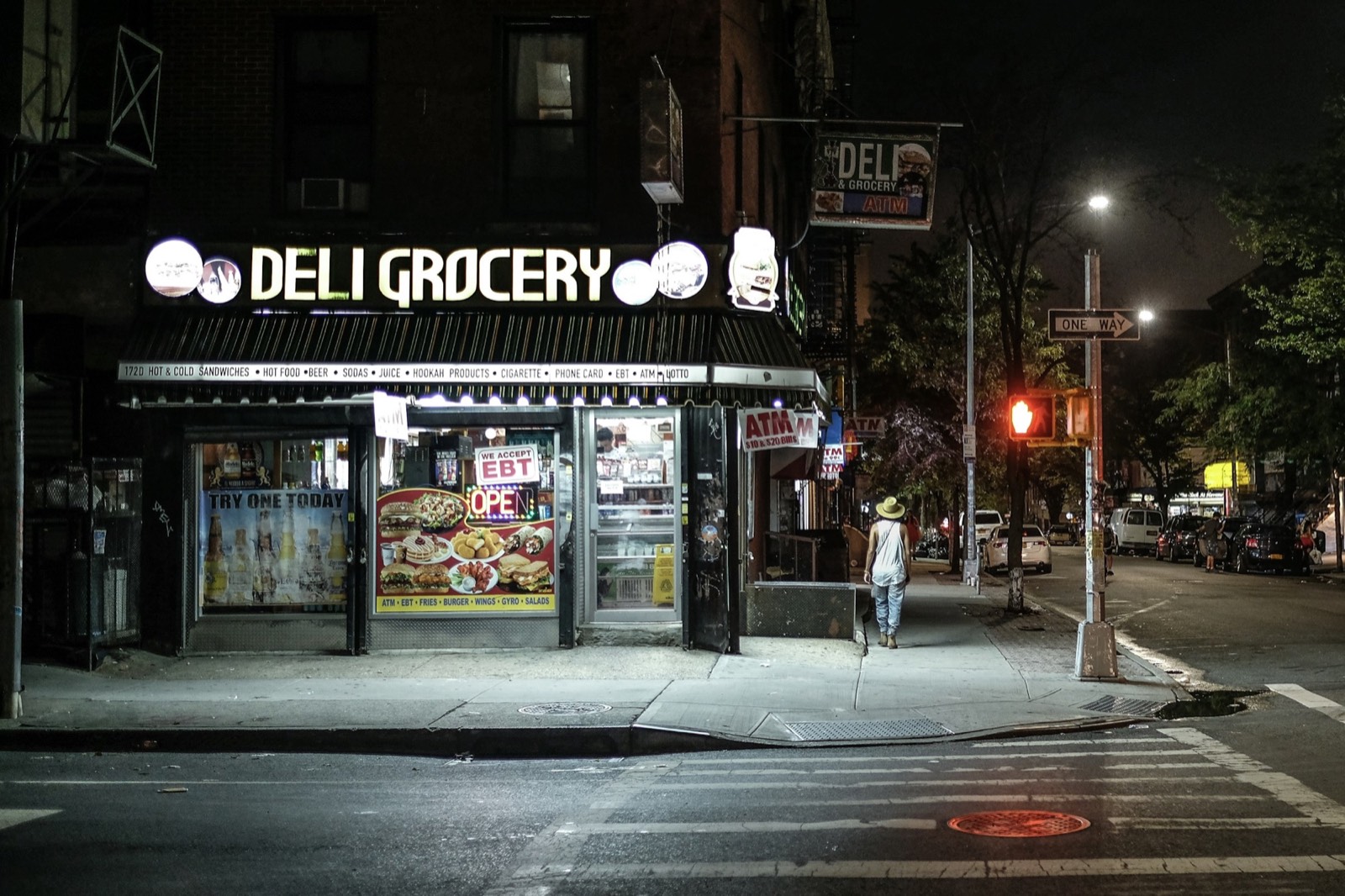 A lit corner deli grocery at night with one person crossing the empty street