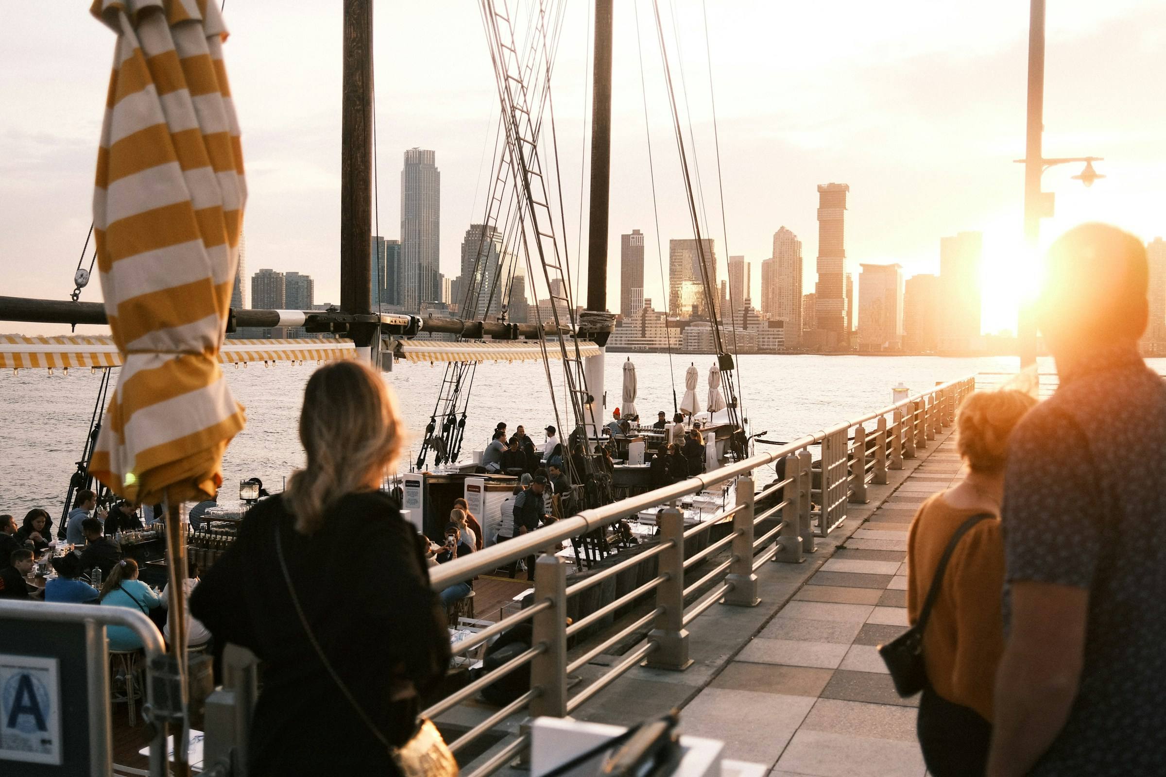People on the Hudson River pier beside the Grand Banks schooner at sunset, with the Jersey City skyline across the water