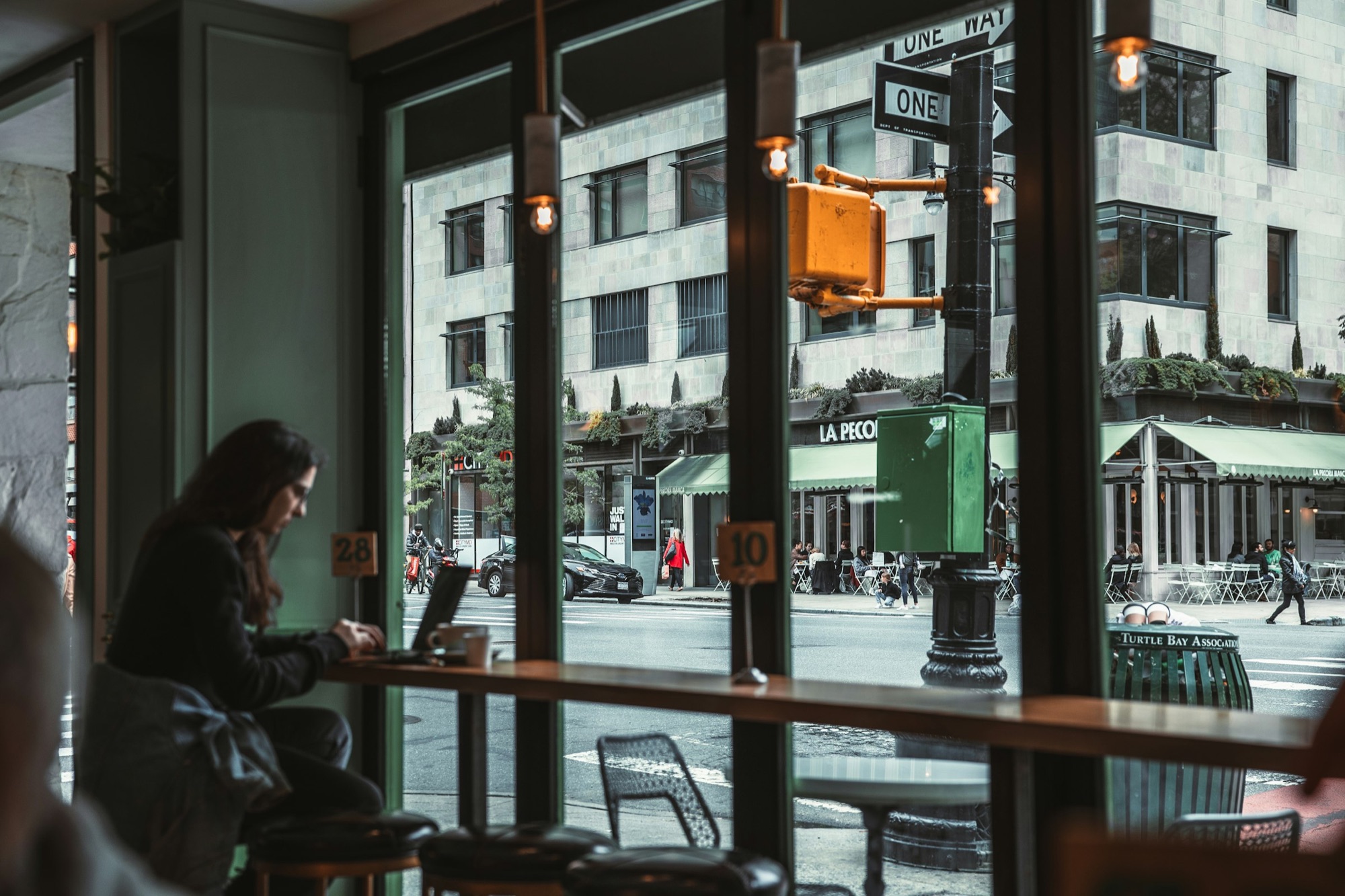 A woman works on a laptop at a long cafe window counter above a Manhattan street corner